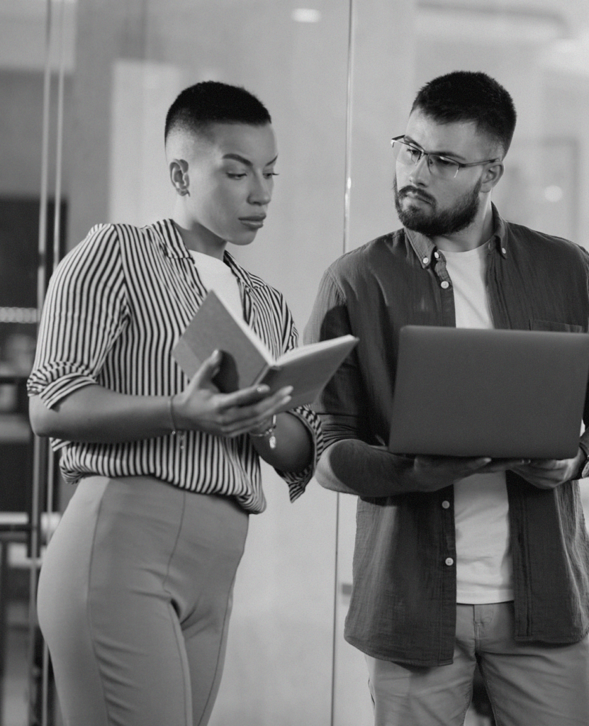 Staff members, one female and one male, discussing notes while standing outside of a conference room.