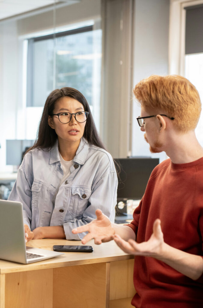 Female and male team members talking in front of a desk that has a laptop and mobile phone on it.